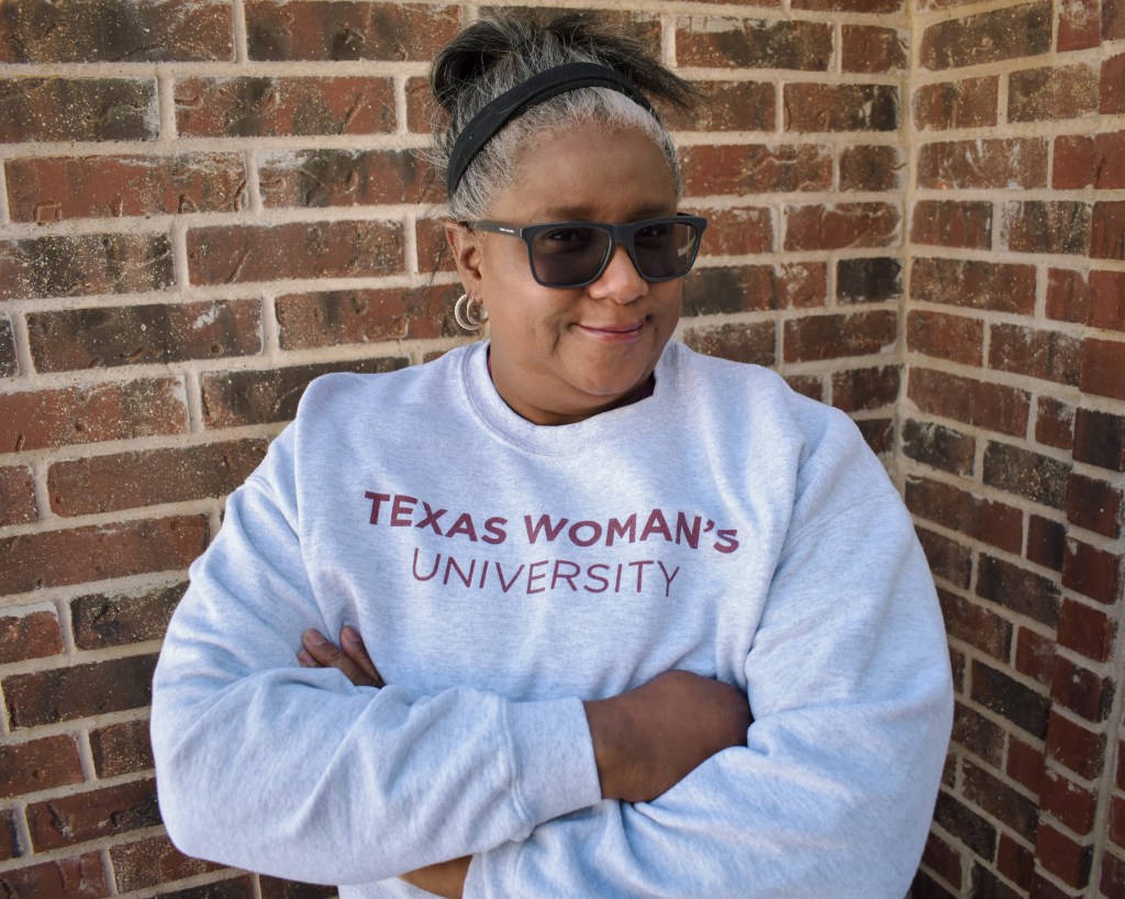 Vicki Byrd stands with her arms crossed and a wise smile on her face in front of some red brick while wearing a light gray sweater with the words Texas Woman's University in maroon